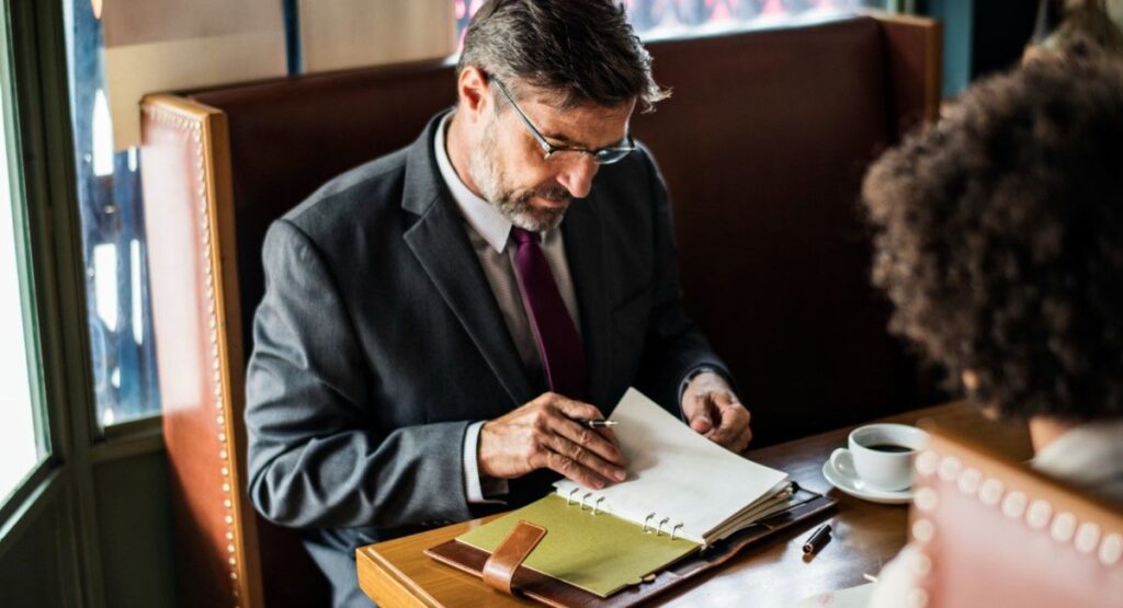 Homem de terno lendo um caderno em um ambiente de cafeteria, simbolizando trabalho ou estudo em um local público