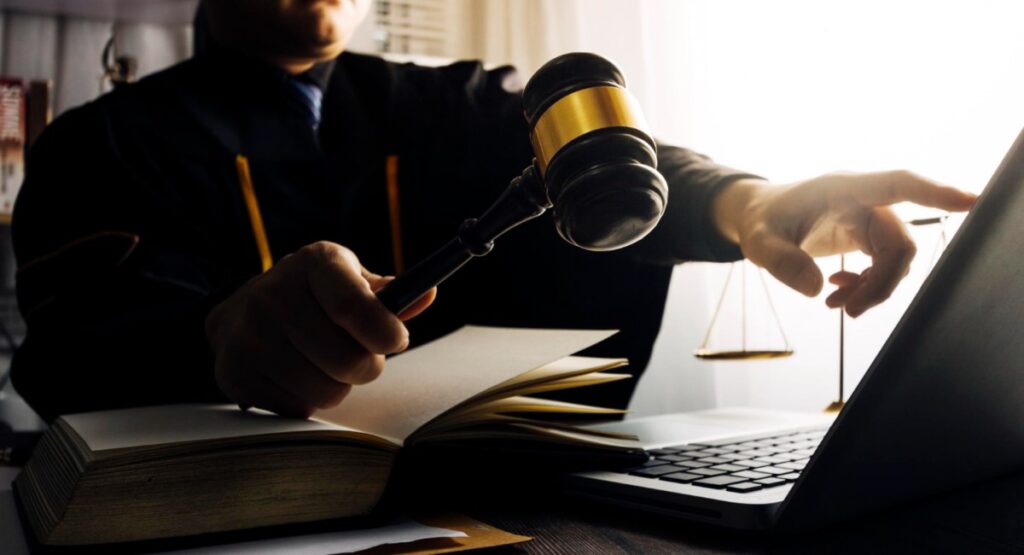 Juiz segurando um martelo enquanto trabalha em um computador, com livros de leis e uma balança da justiça ao fundo, simbolizando o processo judicial e a tomada de decisões legais.