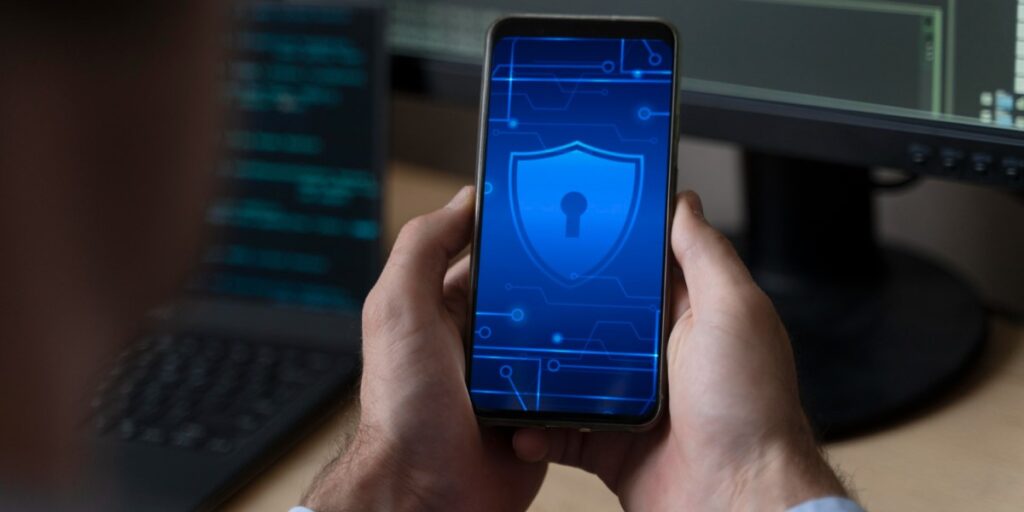 Pessoa segurando um smartphone com um ícone de segurança digital na tela, representado por um escudo com um cadeado. Ao fundo, há um laptop e um monitor exibindo código de programação.