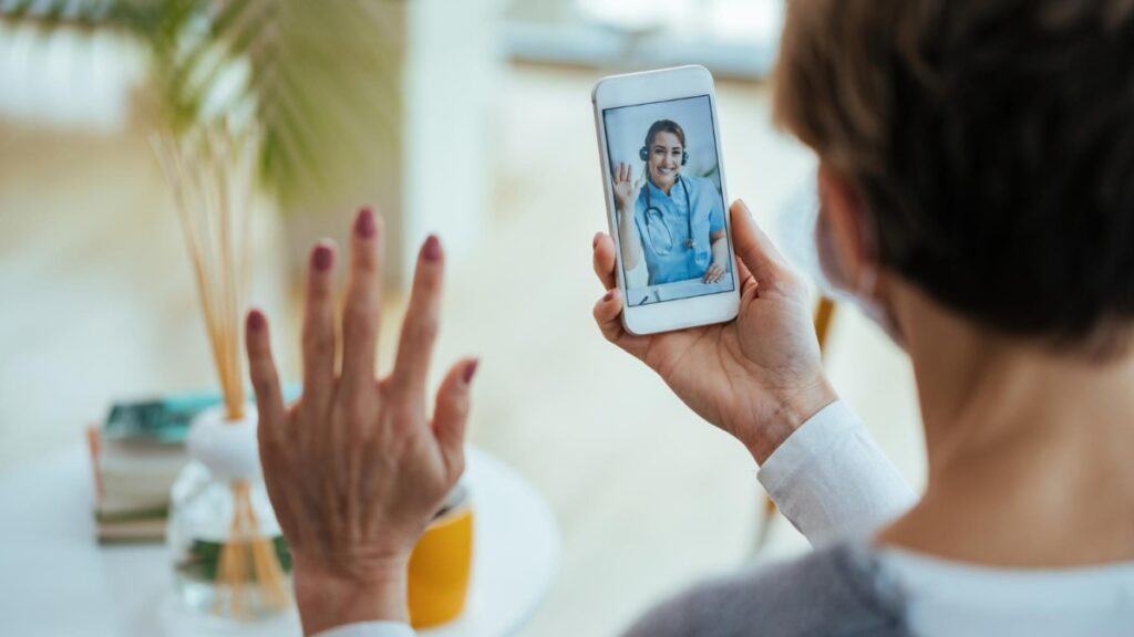 Pessoa acenando com a mão durante uma consulta médica por videoconferência no celular, com a imagem de uma médica na tela, em um ambiente doméstico com plantas ao fundo.
