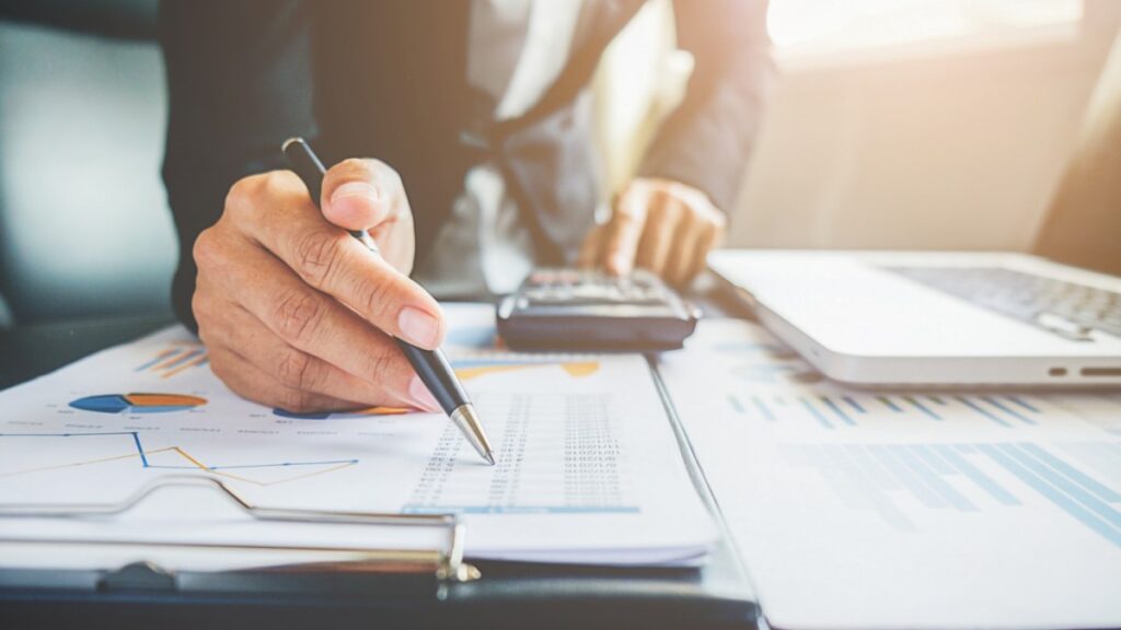 Pessoa de negócios analisando documentos financeiros em uma mesa, com um laptop e uma calculadora ao lado, simbolizando a revisão de finanças ou contabilidade.
