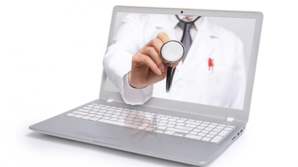 Médico com jaleco branco e estetoscópio segurando um estetoscópio em direção à câmera, aparecendo na tela de um laptop em um conceito de telemedicina.