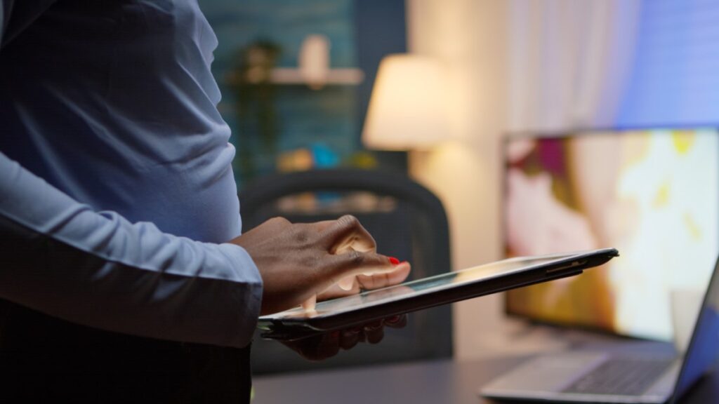 Pessoa usando um tablet em um ambiente de trabalho, com foco nas mãos interagindo com o dispositivo e uma sala iluminada ao fundo.