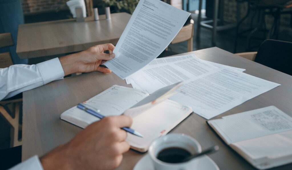 Homem revisa documentos em uma mesa de escritório, com uma xícara de café, um caderno e outros papéis espalhados.