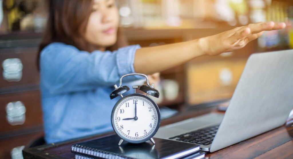 Mulher esticando o braço em direção ao laptop com um despertador analógico na frente, representando a importância da gestão do tempo no trabalho.