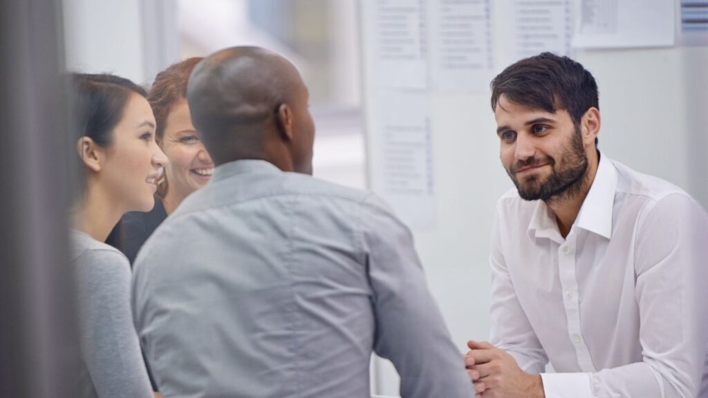 Um grupo de quatro pessoas reunidas em um ambiente de trabalho. Três delas estão de costas, enquanto uma está de frente, vestindo uma camisa social branca e exibindo um sorriso. Eles parecem estar envolvidos em uma conversa amigável.