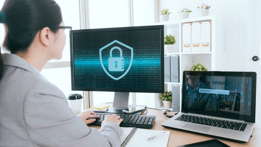 Pessoa usando um computador em um ambiente de escritório, com um monitor exibindo um ícone de cadeado, representando segurança cibernética. Um laptop ao lado também mostra uma tela de login.