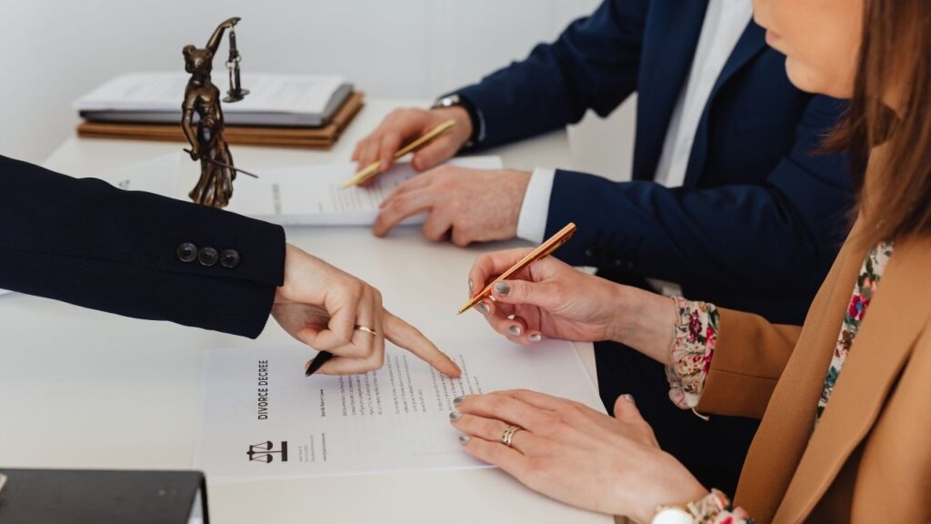 Duas pessoas assinando um documento jurídico em um escritório, com uma estátua da Justiça ao fundo, representando um contrato ou acordo formal sendo firmado.