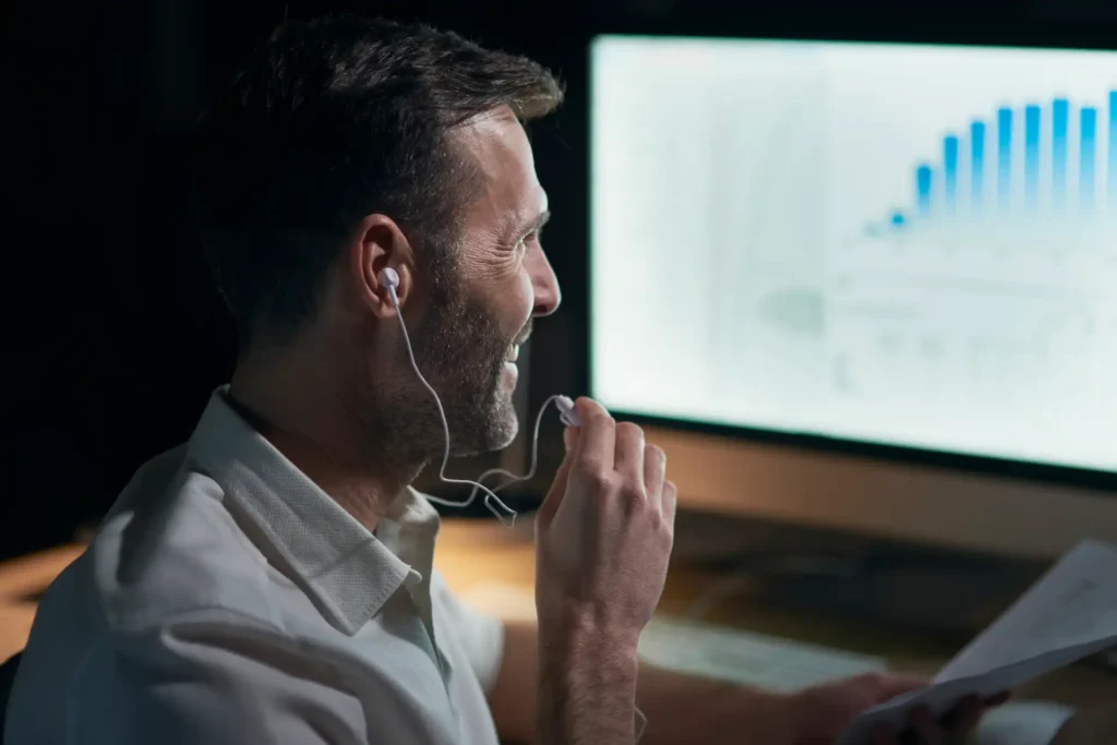 Homem sorridente com fones de ouvido, analisando gráficos em um monitor enquanto segura anotações, representando trabalho e produtividade.