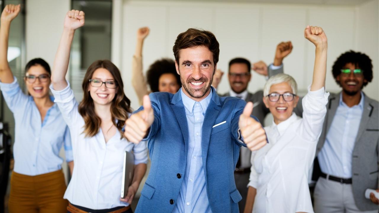 Um homem de terno azul sorridente, fazendo um sinal de positivo com as duas mãos, cercado por uma equipe diversificada e animada ao fundo, transmitindo sucesso, motivação e espírito de equipe.