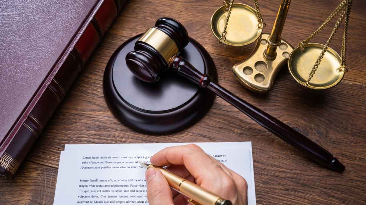 Imagem de um martelo de juiz, balança da justiça e uma mão segurando uma caneta em cima de um documento, simbolizando o direito e a advocacia.