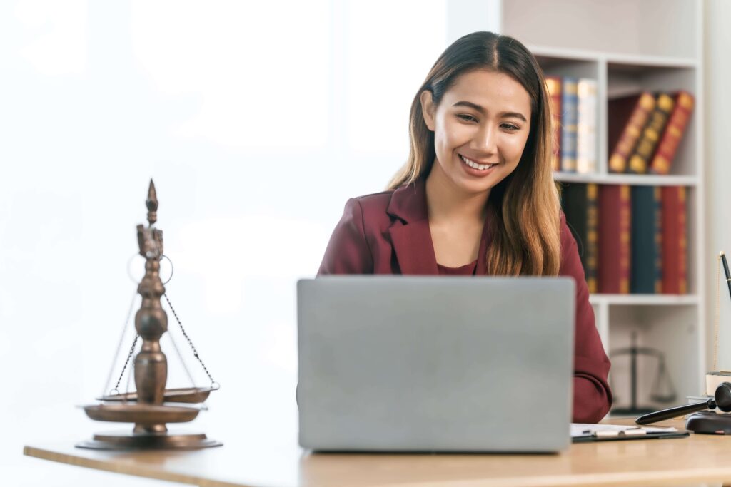 Mulher jovem asiática com cabelo castanho e um blazer bordô, sorrindo enquanto olha para um laptop prateado. Ela está sentada em uma mesa de madeira clara, com uma balança da justiça de bronze no lado esquerdo da imagem e um livro de capa preta e um martelo de juiz (gavel) no lado direito. Ao fundo, uma estante branca cheia de livros coloridos, predominantemente vermelhos e azuis, e uma parede branca.