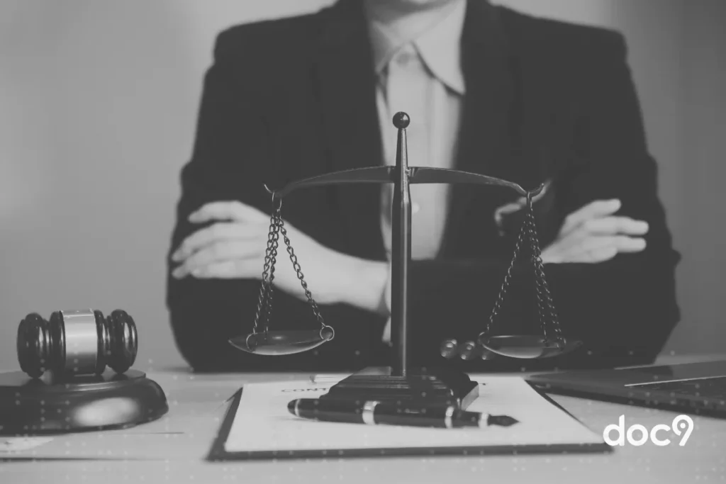 Balança da justiça, martelo de juiz e documentos sobre mesa com pessoa ao fundo vestindo terno, representando uma gestão de departamento jurídico da doc9.